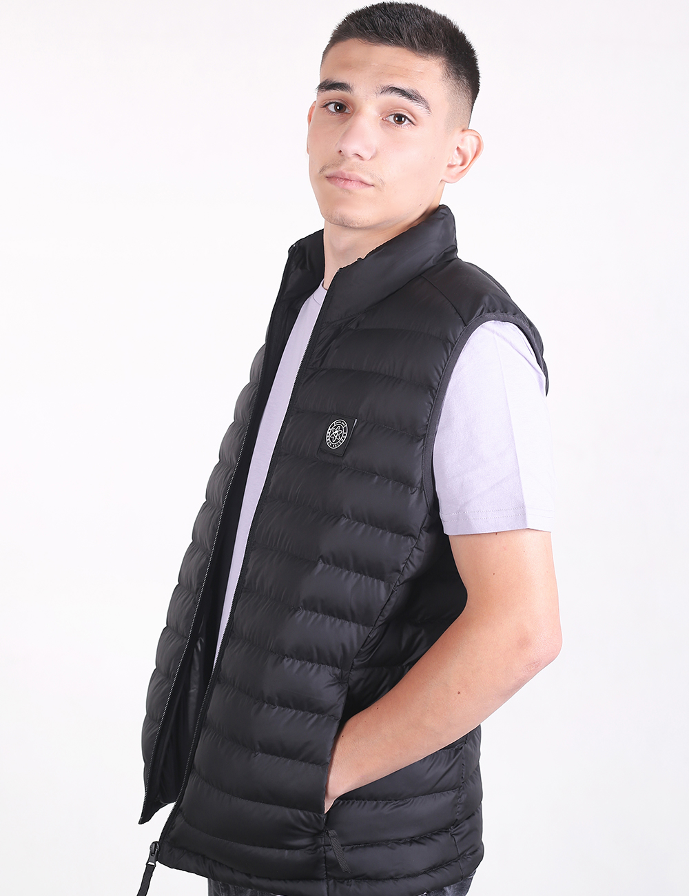 A young man looking toward the camera while posing in a black quilted puffer vest over a light gray t-shirt against a plain white background.