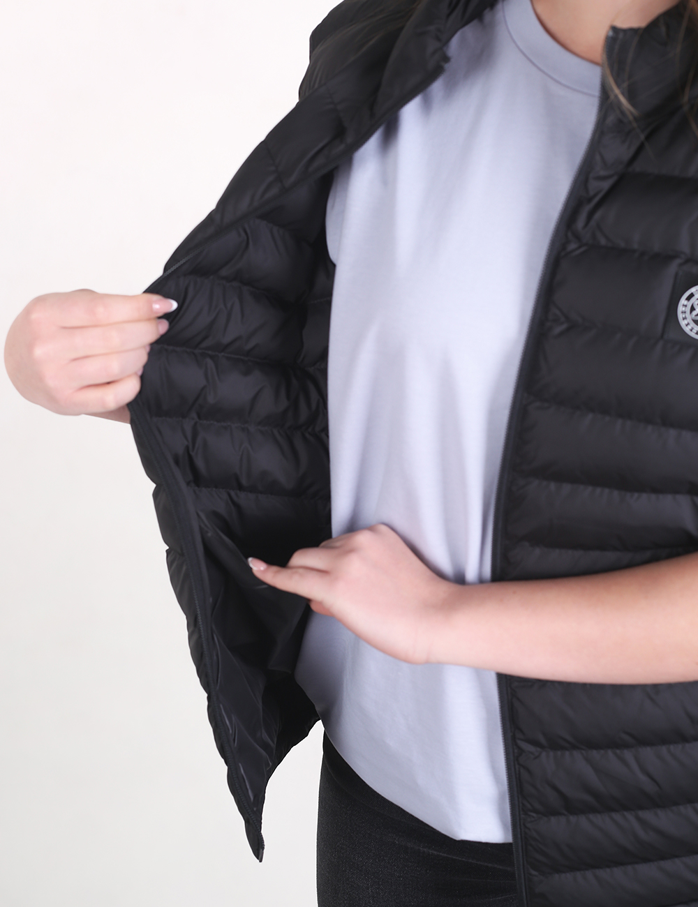 A close-up shot focuses on a person wearing a black quilted puffer vest over a light grey t-shirt and black pants. The individual uses both hands to pull back the right side of the vest, displaying its lightweight design and the matching black interior lining. The vest features thin, horizontal stitched panels and a small, circular white logo on the upper left chest.