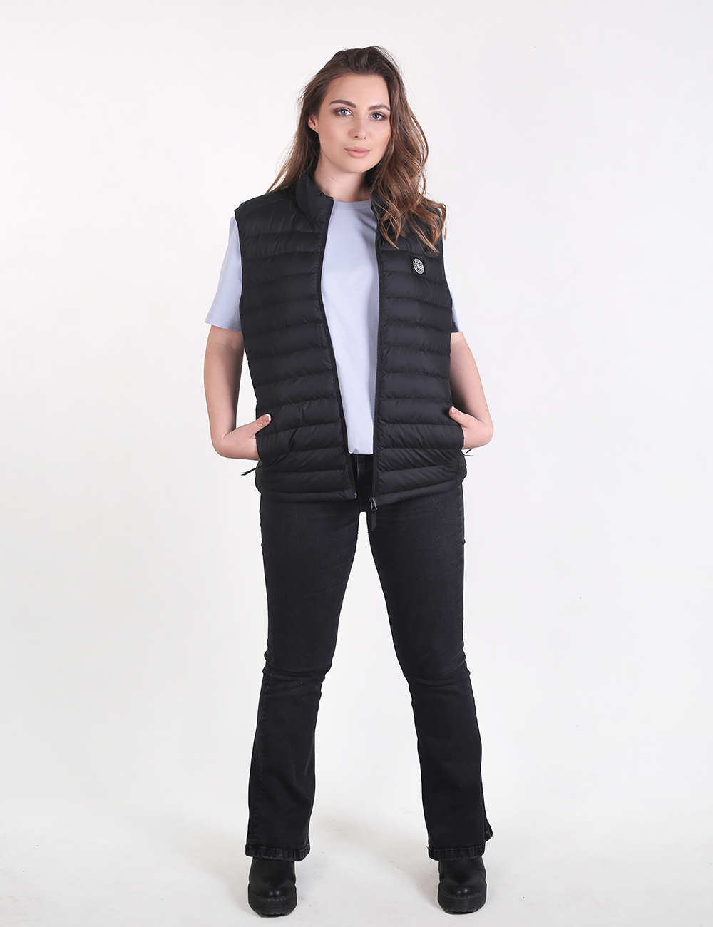A woman with long wavy brown hair stands facing forward against a plain white background, wearing a light purple short-sleeved t-shirt under an open, black quilted puffer vest with a circular logo patch on the left chest. She has her hands in the pockets of the vest and is wearing fitted black pants and black platform shoes.