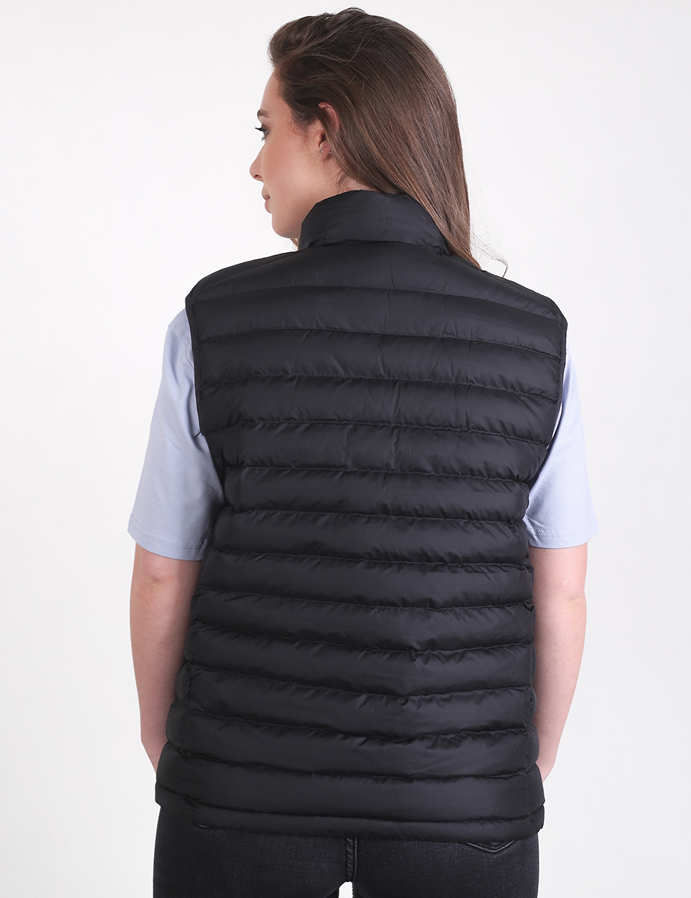 A woman viewed from the back, wearing a black quilted puffer vest over a light-colored, short-sleeved top and dark pants.