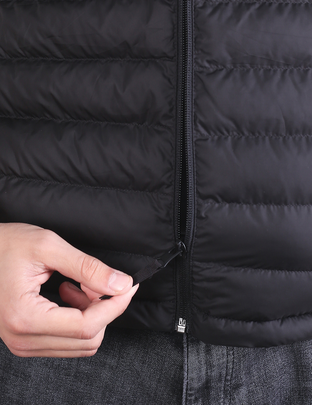 A close-up shot of a person zipping up a black padded puffer vest. The vest features horizontal quilting and a square black patch on the left chest with a circular white atom logo inside.
