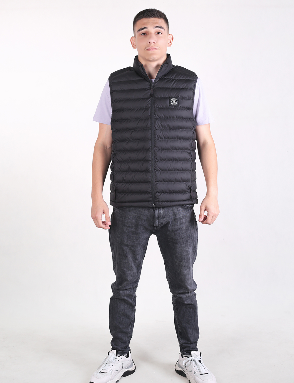 A young man with short dark hair stands facing forward against a plain white background. He is wearing a black, zip-up, quilted puffer vest over a light lavender short-sleeved t-shirt. He is also wearing dark gray slim-fit jeans and chunky white sneakers with black accents. His hands are relaxed at his sides.