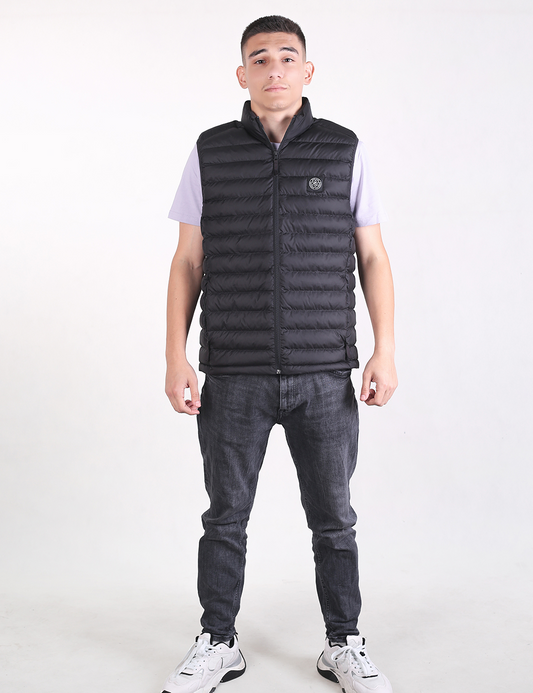 A young man with short dark hair stands facing forward against a plain white background. He is wearing a black, zip-up, quilted puffer vest over a light lavender short-sleeved t-shirt. He is also wearing dark gray slim-fit jeans and chunky white sneakers with black accents. His hands are relaxed at his sides.