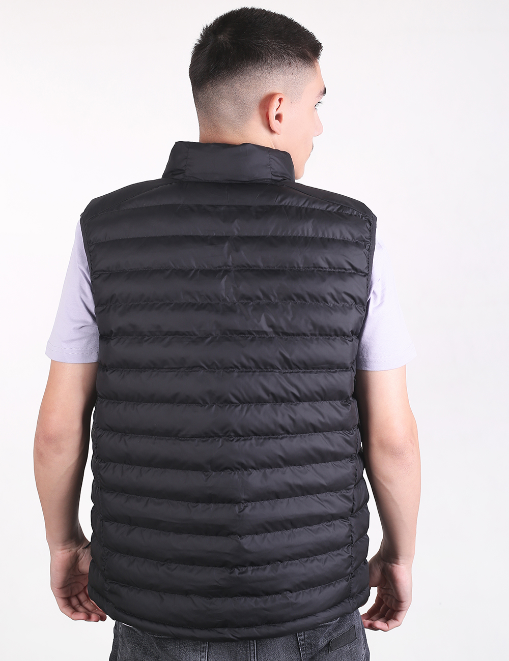 Rear view of a man wearing a black, quilted puffer vest over a short-sleeved, light grey t-shirt. The vest features horizontal stitching and a stand-up collar.