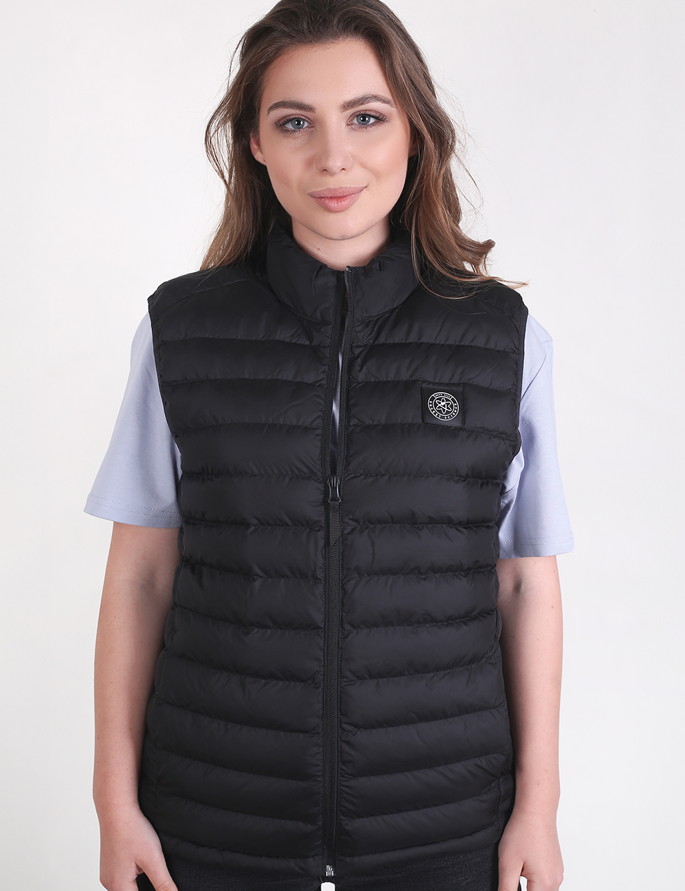 A woman with long brown hair posing against a plain light gray background, wearing a black puffer vest over a light-colored short-sleeved t-shirt.