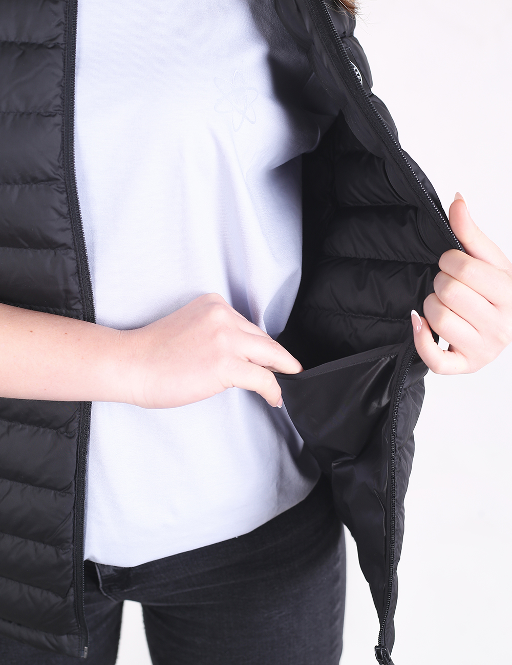 A close-up shot focuses on a person wearing an open black quilted puffer vest over a plain light grey t-shirt. The vest features narrow horizontal stitching and a smooth inner lining. The person’s right hand gently pulls open the side of the vest, while their left hand is tucked inside a secure, zippered side pocket, highlighting its functionality. The background is a clean, neutral white.