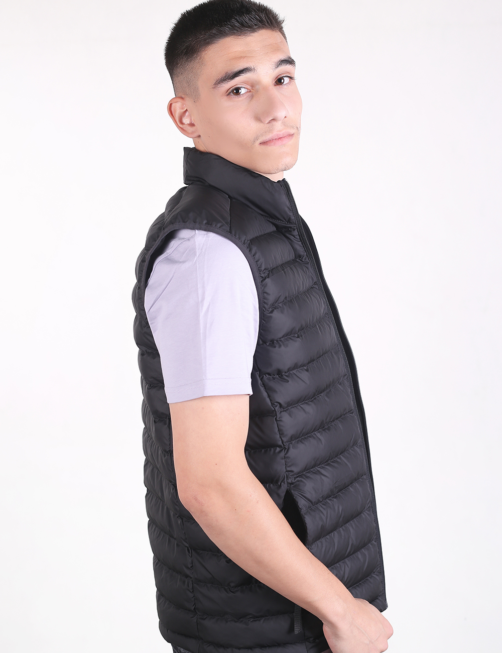 A young man with short dark hair, shown from the side in a profile view against a plain white background, wearing a white short-sleeved t-shirt under a black, lightweight quilted puffer vest with horizontal stitching.