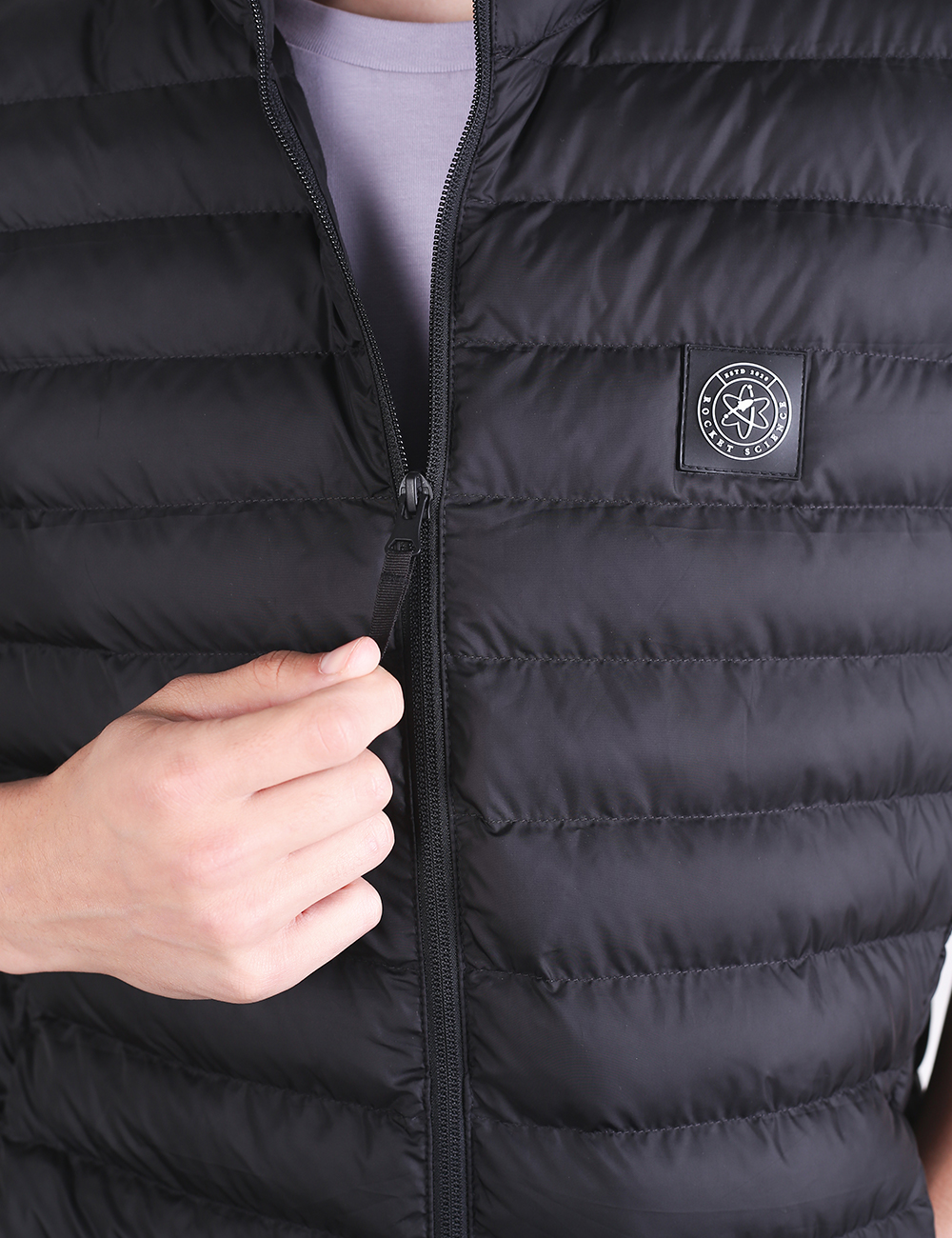 A close-up shot of a person zipping up a black padded puffer vest. The vest features horizontal quilting and a square black patch on the left chest with a circular white atom logo inside.