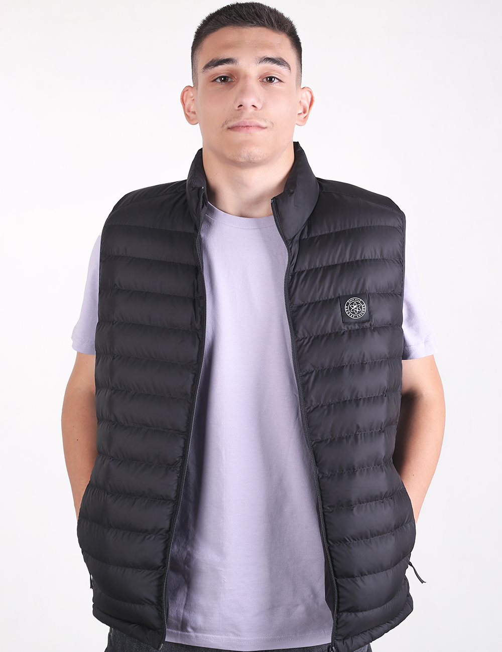 A young man with short dark hair stands looking forward, wearing an open, black quilted puffer vest over a plain, light purple t-shirt. The vest features narrow horizontal baffles and a small, circular logo patch on the left chest. His hands are tucked behind his back or into his pockets against a plain, light grey background.