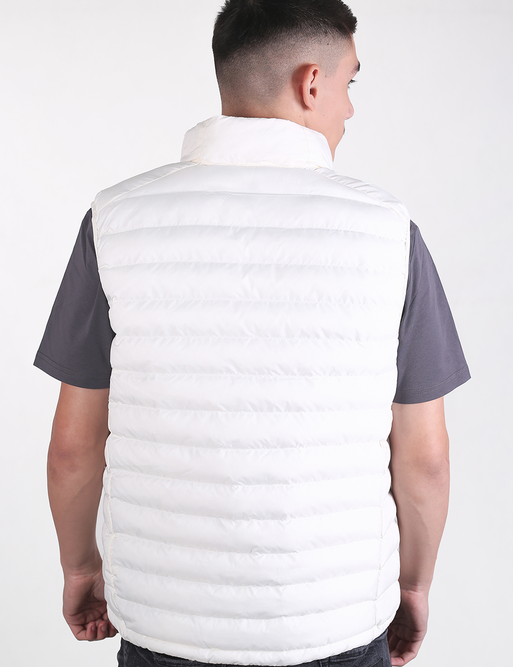 A man viewed from the back wearing a white quilted puffer vest over a dark gray short-sleeve shirt, against a plain light gray studio background.