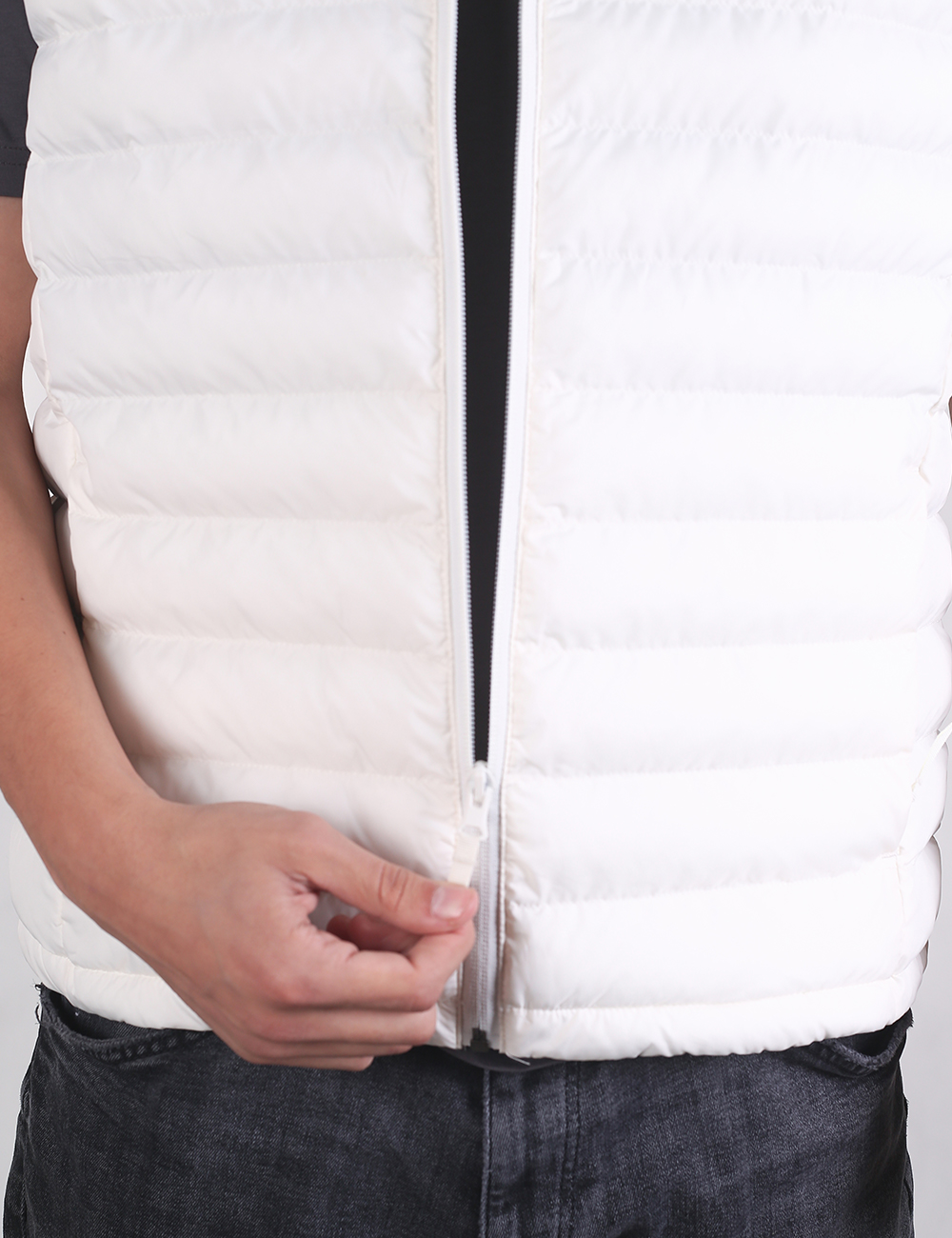 A close-up of a white quilted puffer vest being worn, with a person's hand adjusting the zipper near the hem, paired with dark gray jeans.