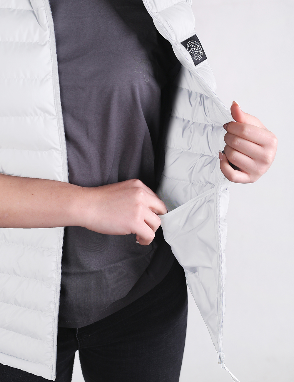 A close-up shot focuses on a person wearing a lightweight, off-white quilted puffer vest open over a charcoal gray t-shirt and black pants. The person's left hand is tucking the inner lining of the vest forward, while their right hand rests near the vest's pocket. A small, circular black brand patch is visible on the upper left chest area of the vest.