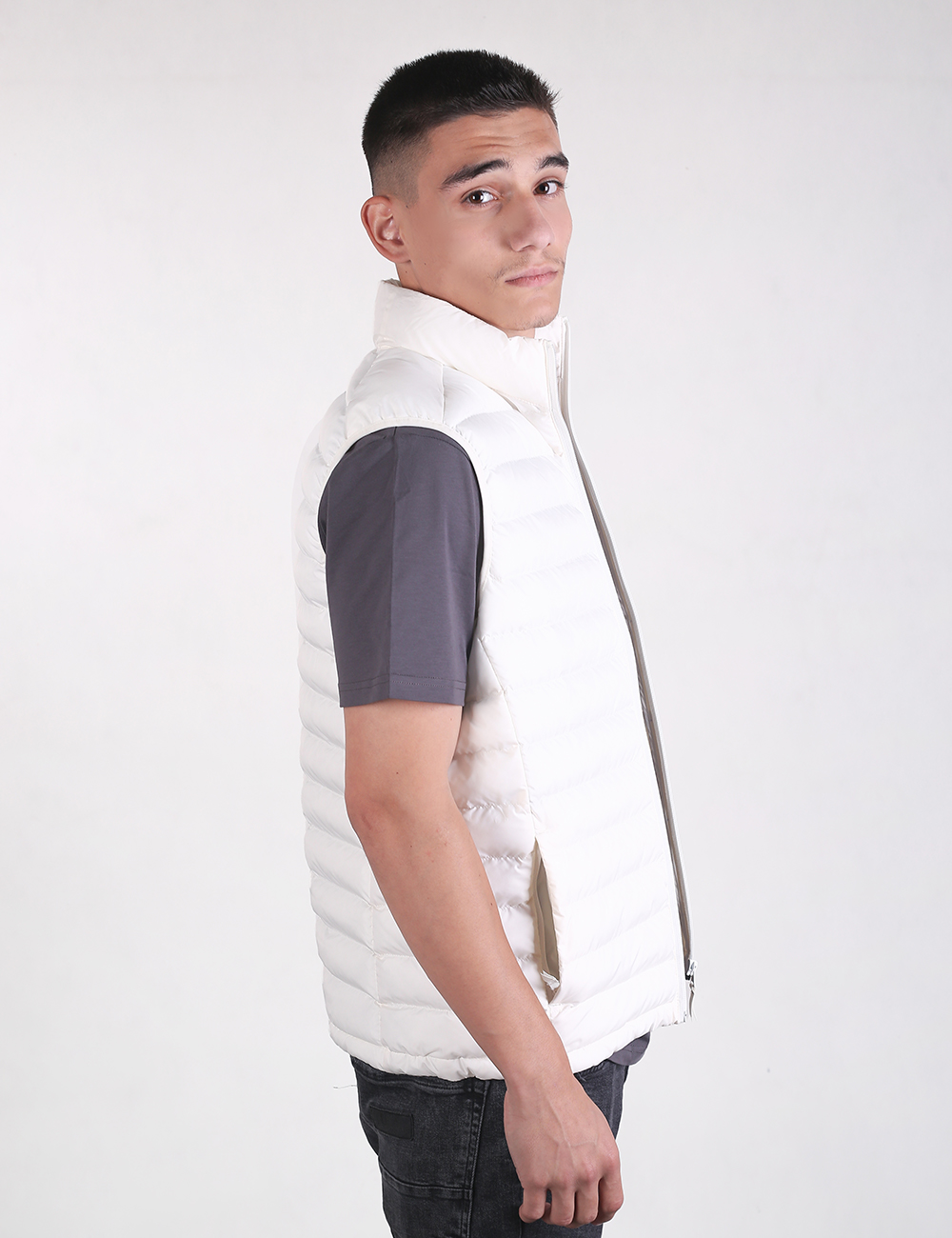A man wearing a white puffer vest over a dark gray short-sleeve shirt, shown from the side against a plain light gray studio background.