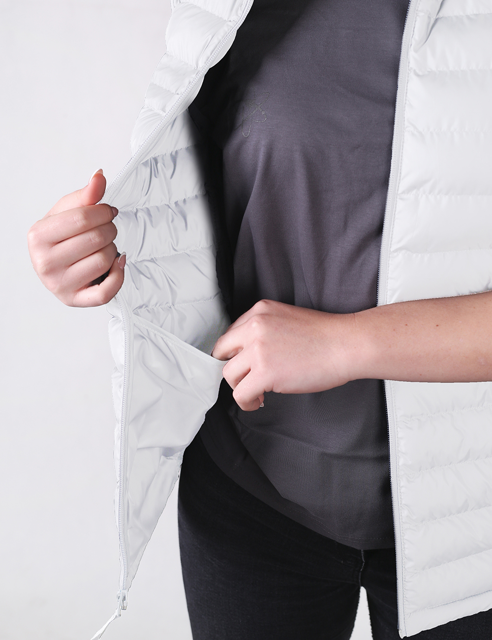 A close-up shot focuses on a person wearing an open, lightweight white puffer vest over a charcoal gray t-shirt and black pants. The person's right hand holds the inner lining of the vest open, revealing a hidden interior pocket where their left hand is tucked inside.