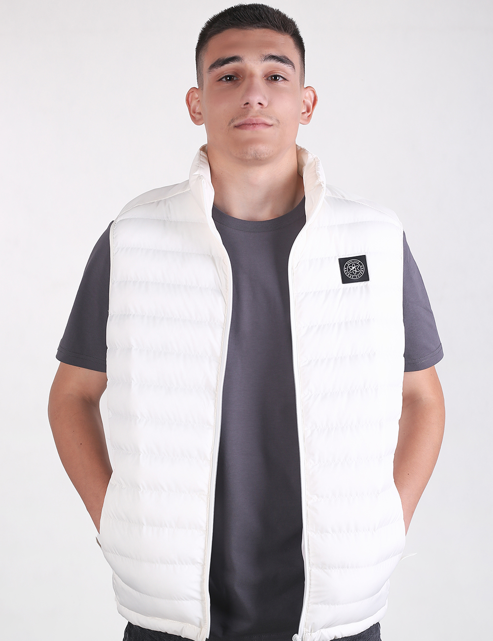 A man wearing a white puffer vest over a dark gray t-shirt, standing against a light gray studio background.
