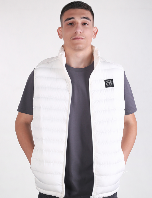 A man wearing a white puffer vest over a dark gray t-shirt, standing against a light gray studio background.