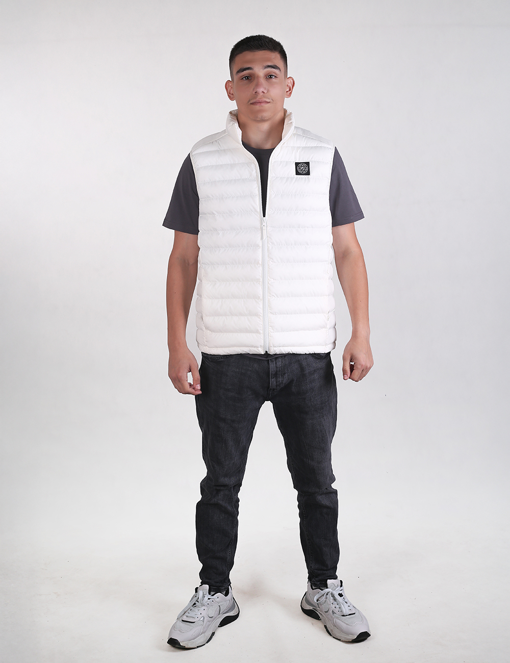 A man modeling a white puffer vest over a dark gray t-shirt, black jeans, and gray sneakers, standing against a light gray studio background.
