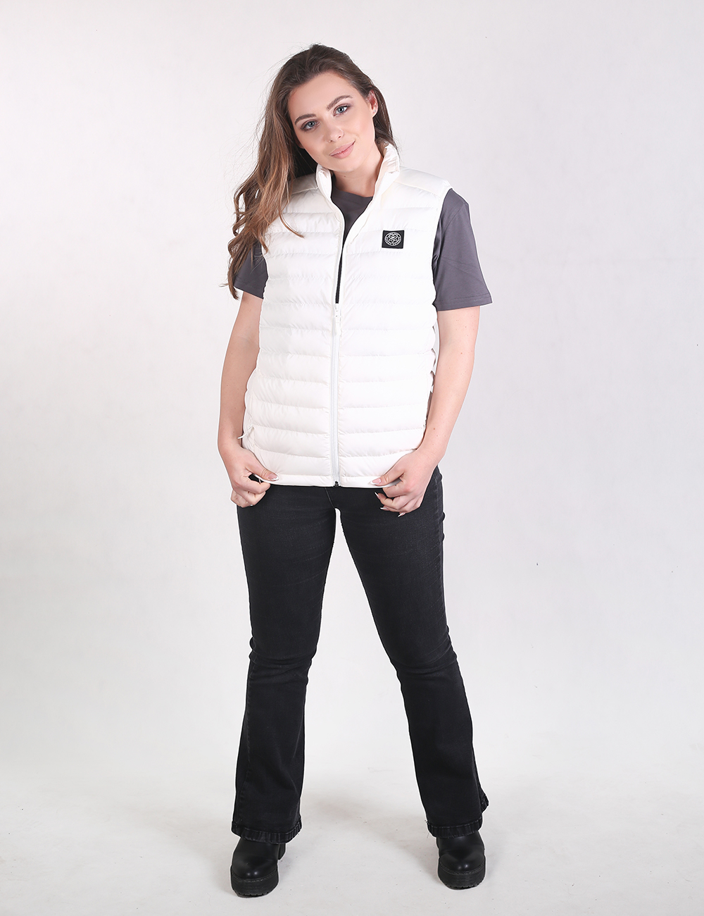 A woman modeling a white sleeveless puffer vest over a dark gray short-sleeve shirt, paired with black pants and black shoes, against a light gray studio background.