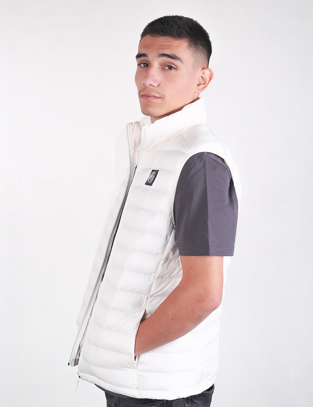 A young man with short dark hair looking over his right shoulder. He is wearing a white puffer vest over a dark grey short-sleeve t-shirt. The vest features horizontal quilting, a high collar, a zipper closure, and a small black rectangular patch on the left chest. He has his hands placed inside the pockets. The background is a solid, light grey color.