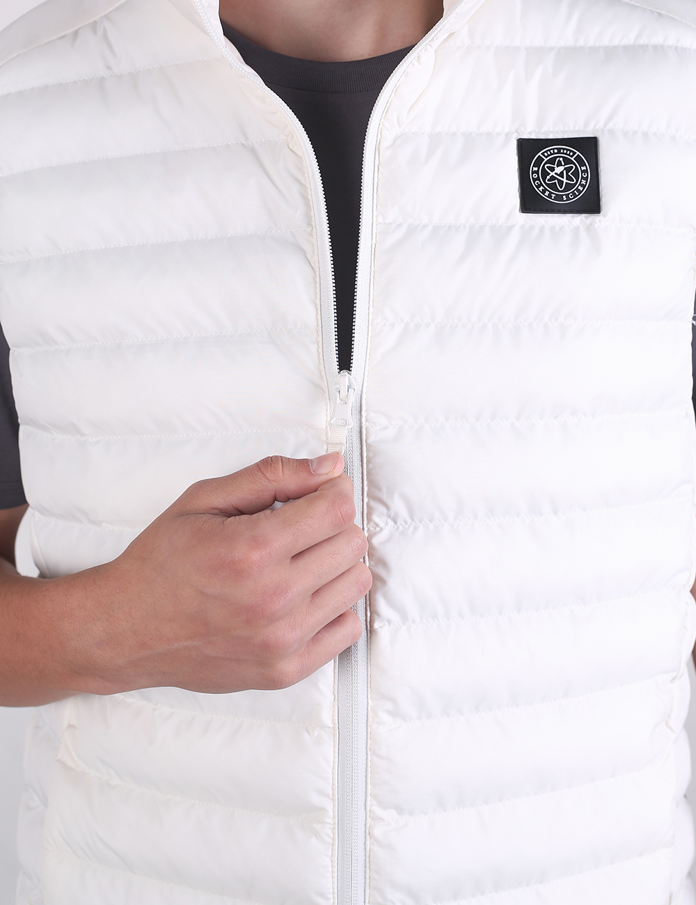 A close-up of a white puffer vest being worn, with a person's hand holding the zipper. The vest features horizontal quilting, a black zipper, and a black square patch on the left chest with a white circular logo.