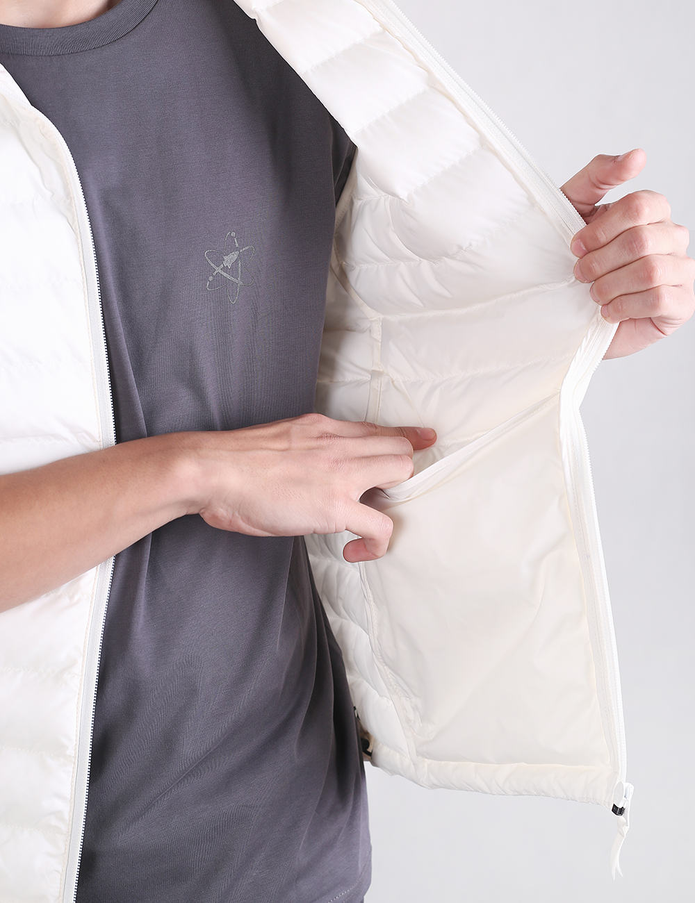 A close-up shot showing a person's torso as they wear a white padded vest over a dark gray t-shirt. The individual's left hand pulls back the front panel of the vest to reveal a deep, built-in interior drop pocket, while their right hand dips into the pocket to demonstrate its depth.