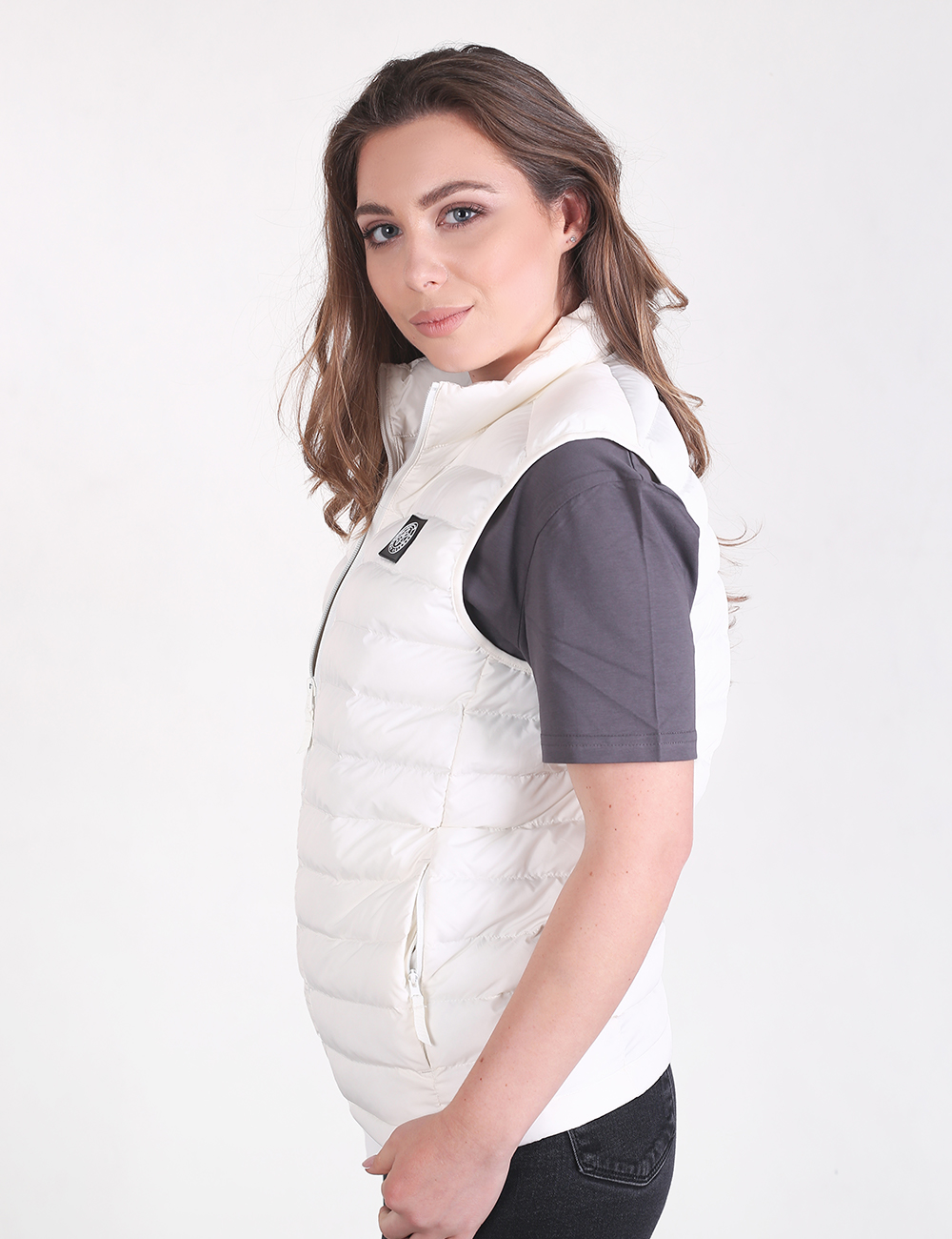 A young woman with long, wavy brown hair looks toward the camera in a side-profile view. She is wearing an off-white, quilted puffer vest over a plain, short-sleeved dark gray t-shirt and black jeans. The vest is zipped up and has a small square logo patch on the upper left chest. The background is a solid, light gray.