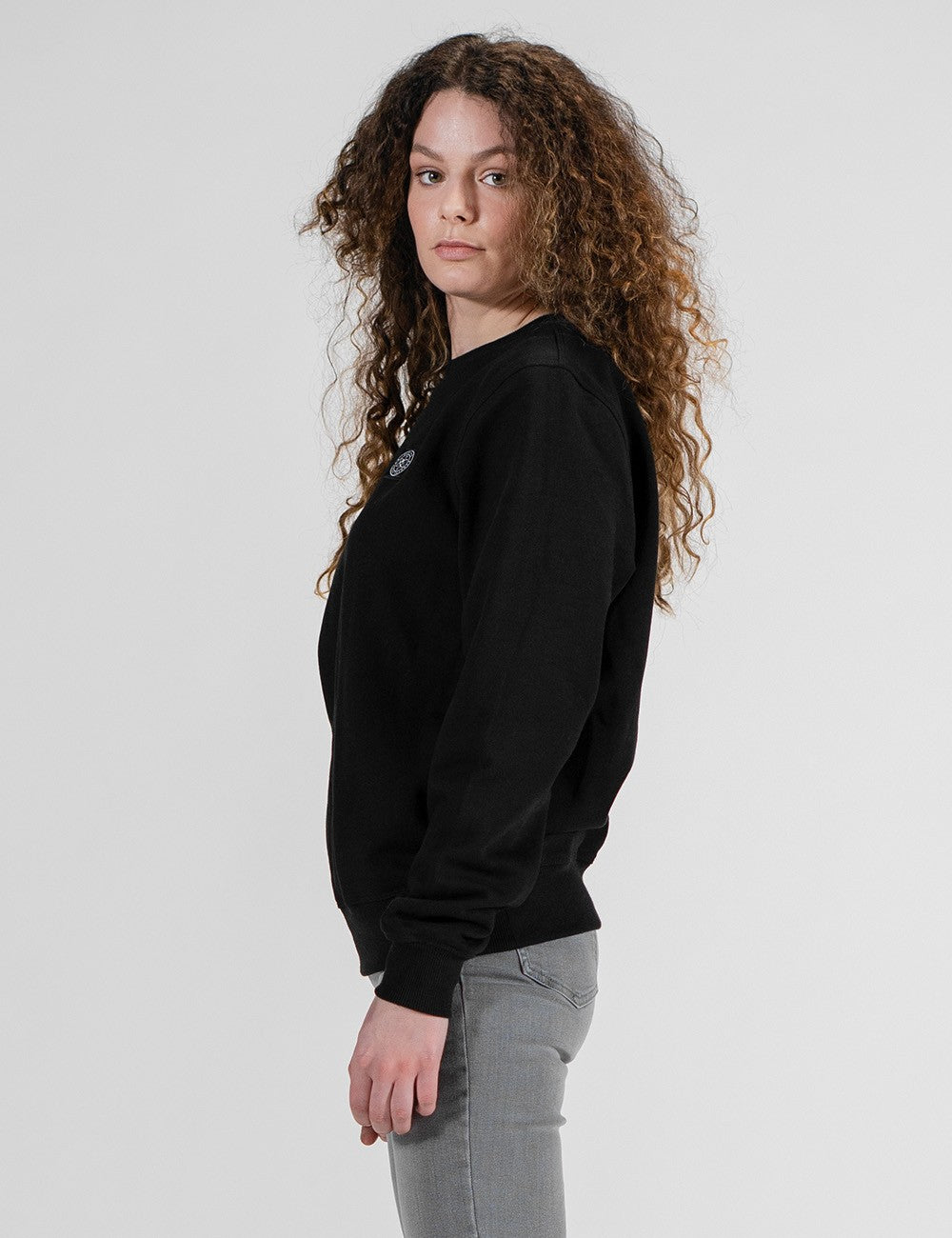 A woman with long, curly brown hair is shown from the side profile, wearing a black crewneck sweatshirt and light gray jeans against a plain light gray studio background.