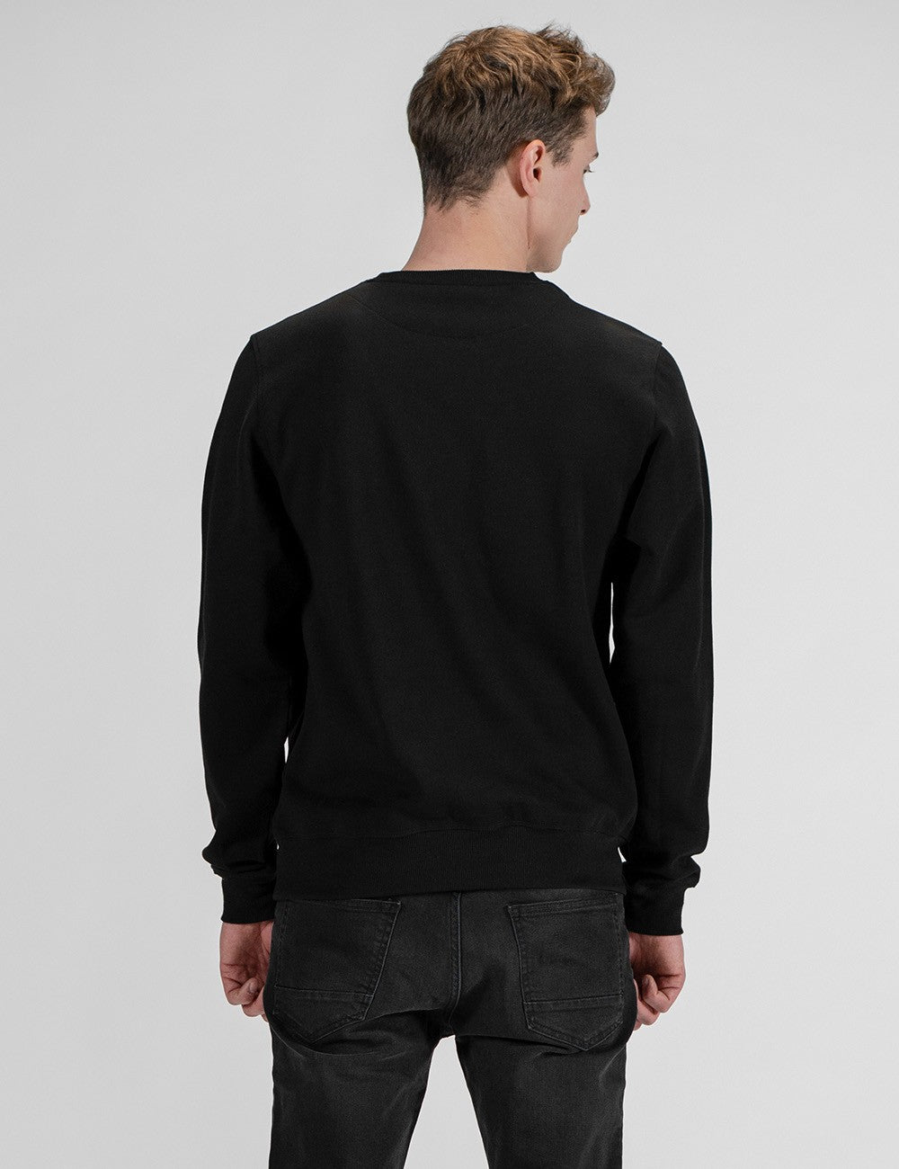 A man viewed from behind wearing a black crewneck sweatshirt and dark gray jeans, standing against a light gray studio background.
