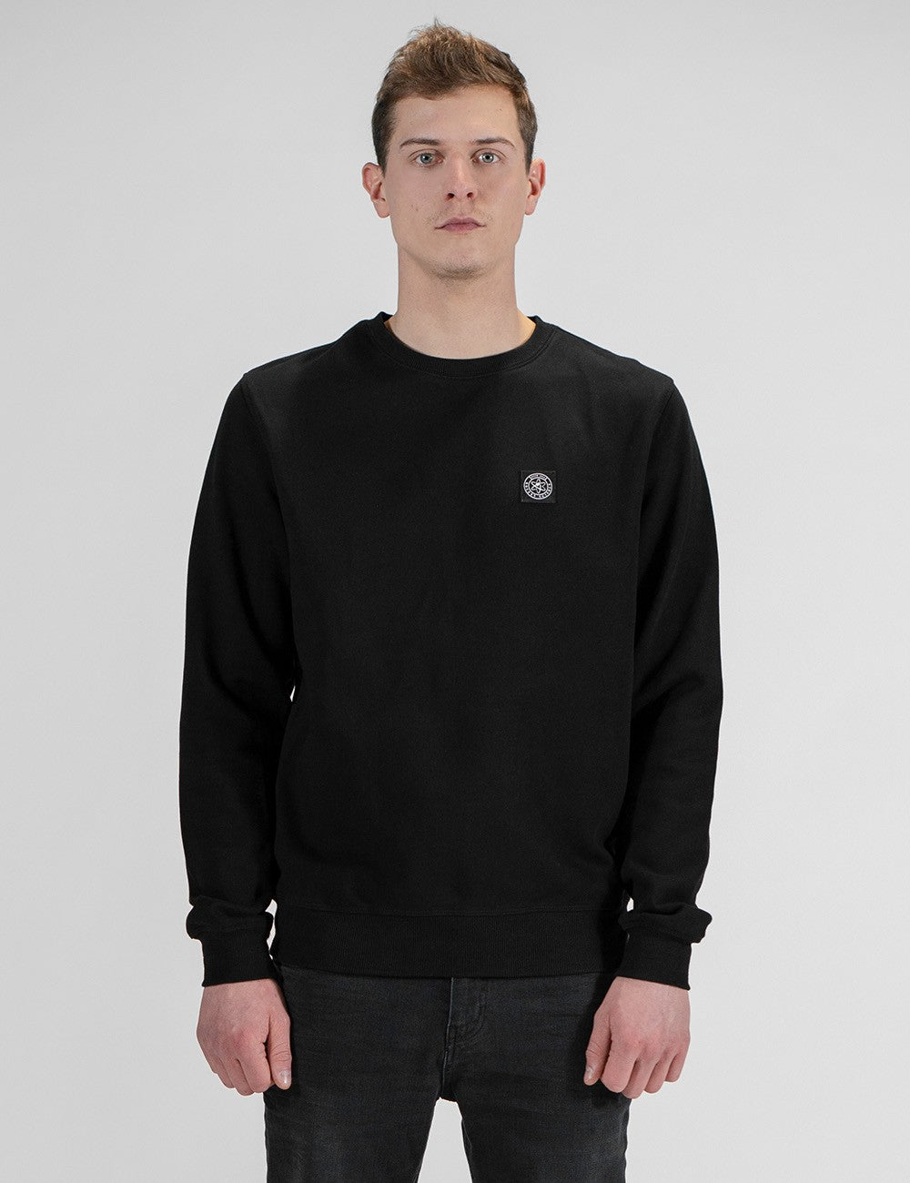 A man wearing a black crewneck sweatshirt with a small circular logo on the left chest, paired with dark jeans, standing against a light gray studio background.