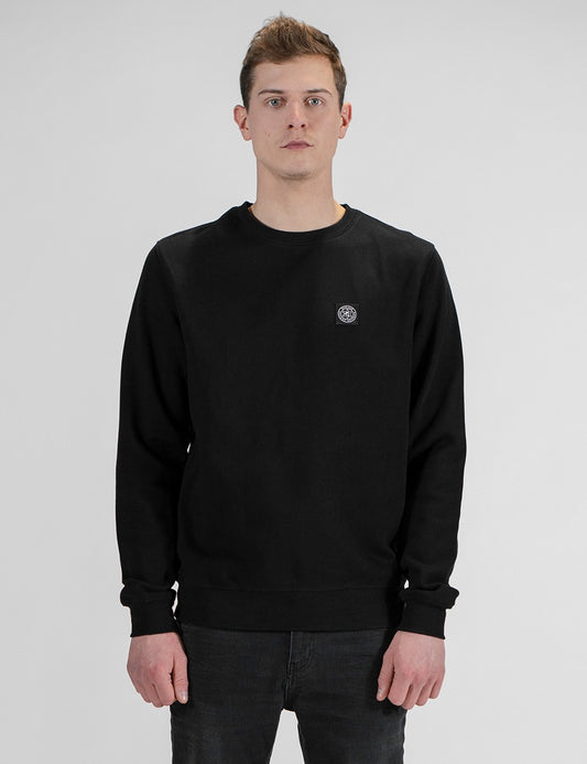 A man wearing a black crewneck sweatshirt with a small circular logo on the left chest, paired with dark jeans, standing against a light gray studio background.