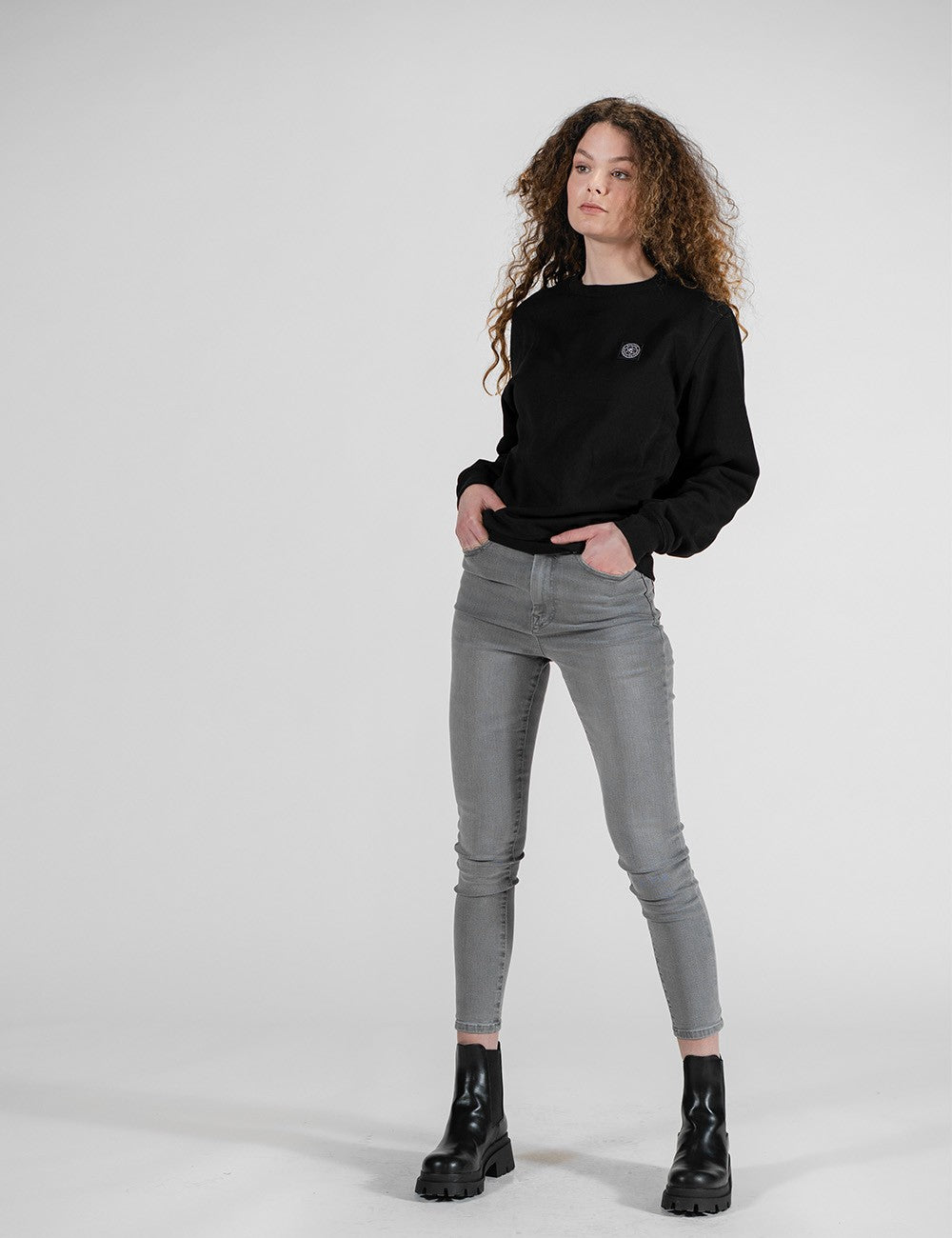 A model with curly brown hair stands against a light gray studio background wearing a black crewneck sweatshirt, gray skinny jeans, and black leather ankle boots.
