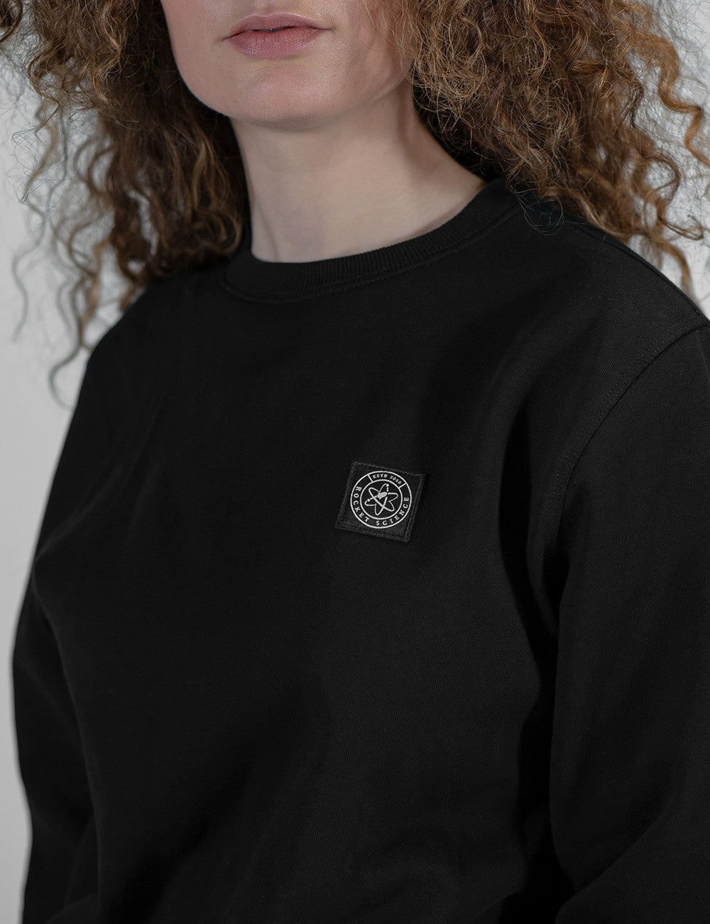 A close-up of a person wearing a black crewneck sweatshirt with a small black-and-white circular logo patch on the left chest. The person has curly light brown hair and is shown from the mouth down to the torso against a plain light background.