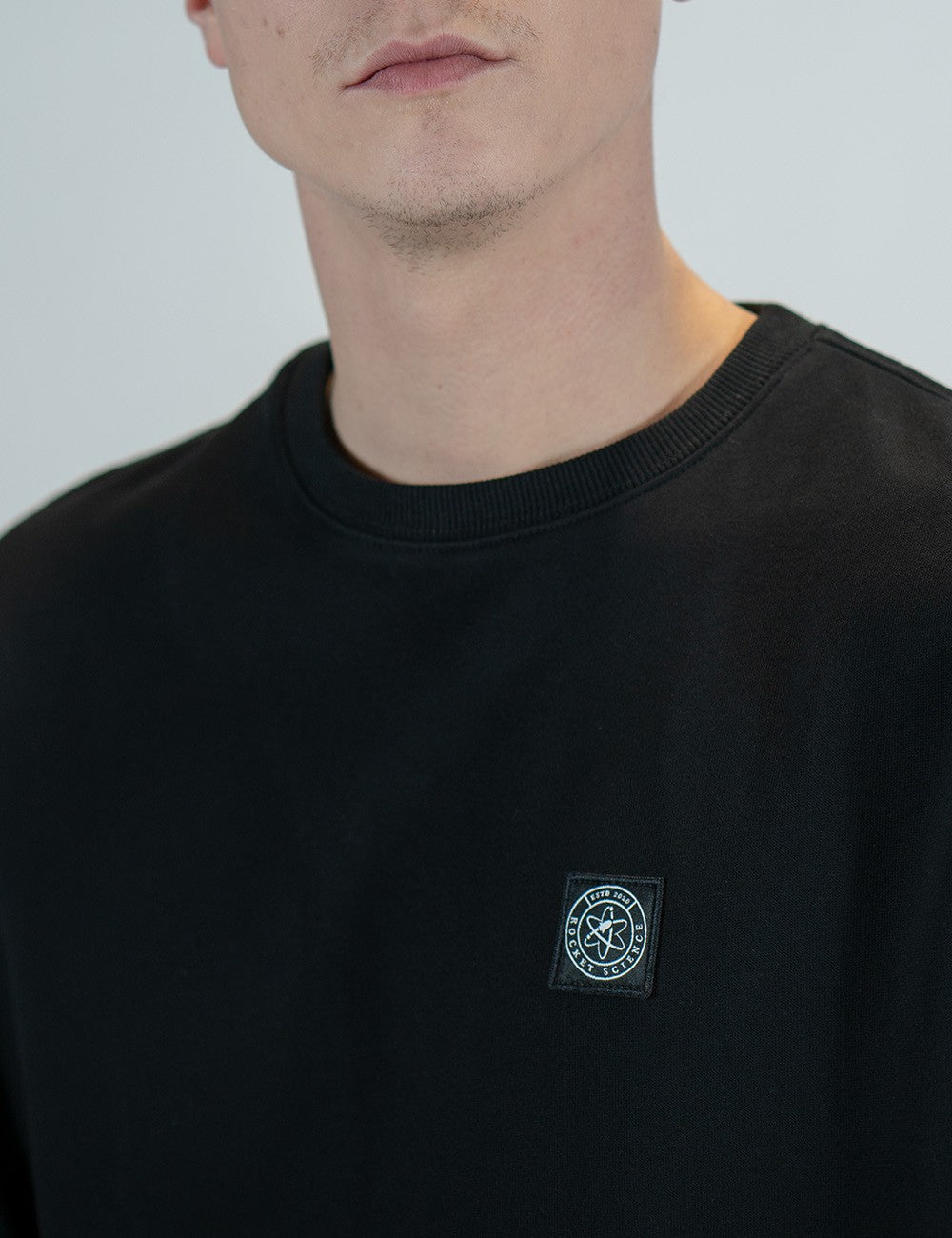 Close-up of a person wearing a black crewneck sweatshirt with a small embroidered  Rocket Science logo on the left chest, against a light gray background.