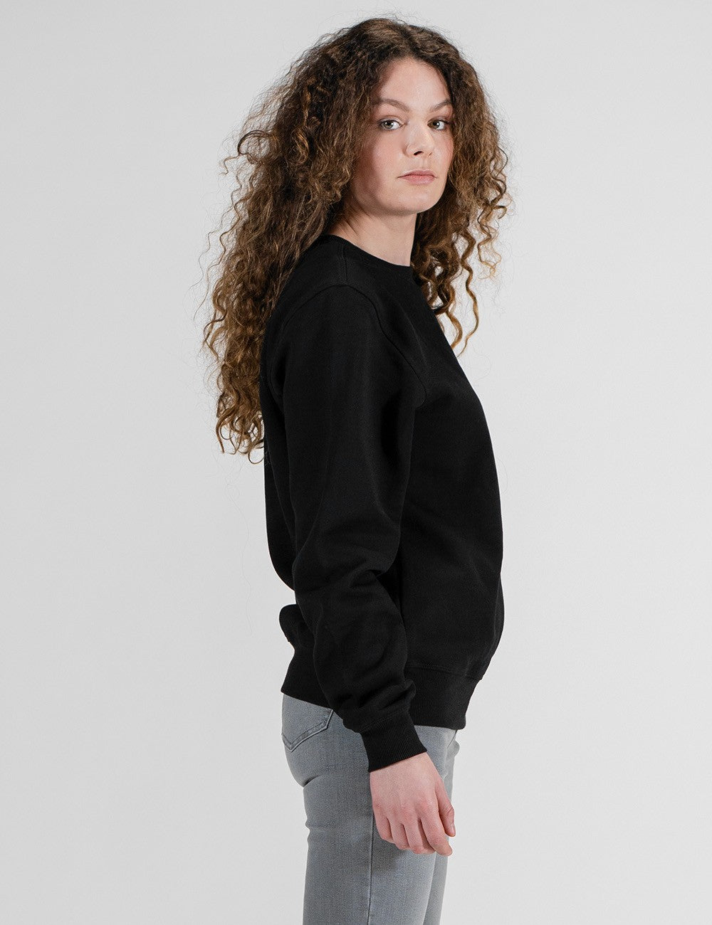 A woman with long, curly brown hair is shown in profile wearing a black crewneck sweatshirt and light gray jeans against a plain light gray studio background.