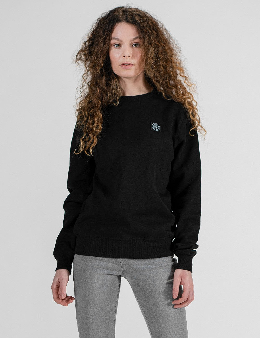 A woman with long, curly brown hair wearing a black crewneck sweatshirt with a small circular logo on the left chest, paired with gray jeans, photographed against a light gray studio background.