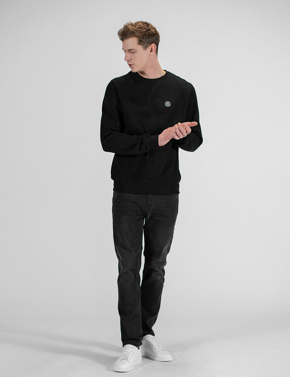 A man wearing a black crewneck sweatshirt, dark gray jeans, and white sneakers, standing against a light gray studio background.