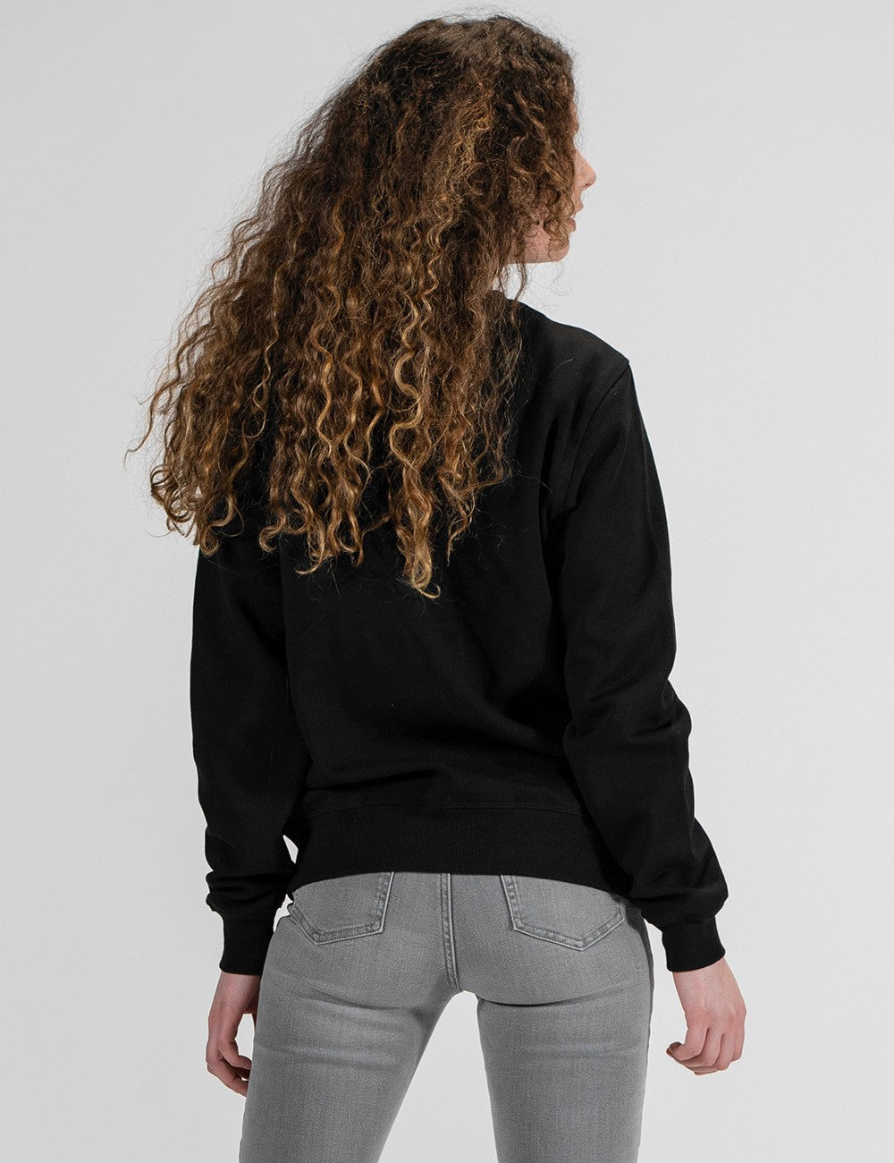 A woman with long, curly brown hair is shown from behind wearing a black crewneck sweatshirt and light gray jeans against a plain light gray studio background.