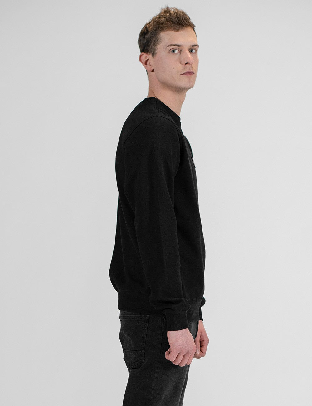 A man in a black crewneck sweatshirt and dark jeans, shown in profile against a light gray studio background.