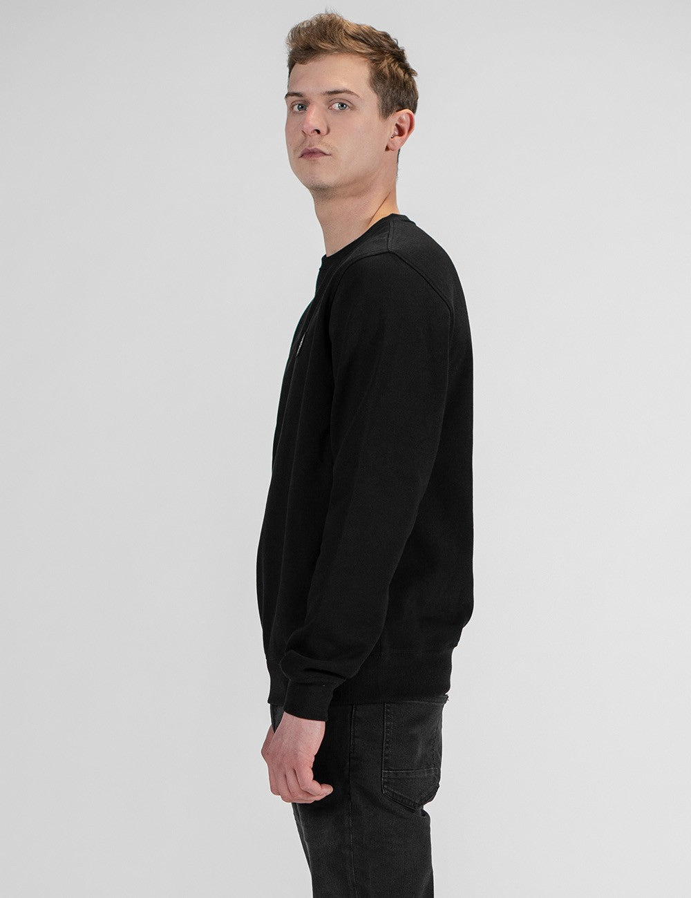 A man in a black crewneck sweatshirt and dark jeans, shown in profile against a light gray studio background.