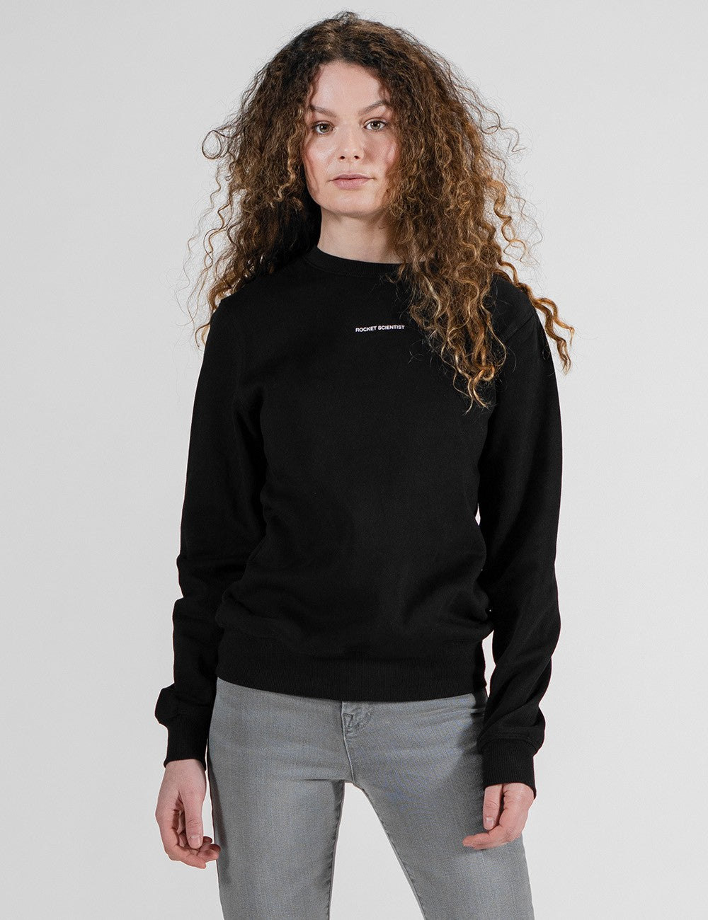 A woman with long, curly brown hair stands facing forward, wearing a black long-sleeved crew-neck sweatshirt featuring a small white text logo on the center chest, paired with grey jeans against a plain light grey background.