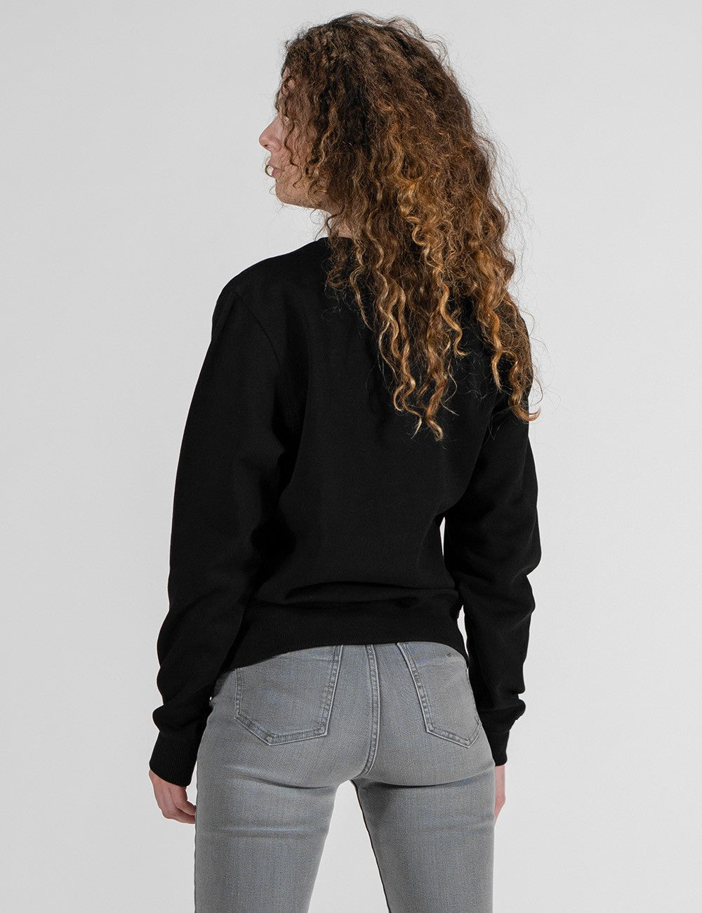 A back-view medium shot of a woman with long, curly blonde and brown hair facing slightly to the left against a solid light gray background. She is wearing a long-sleeved black crewneck sweatshirt and tight-fitting gray denim jeans.