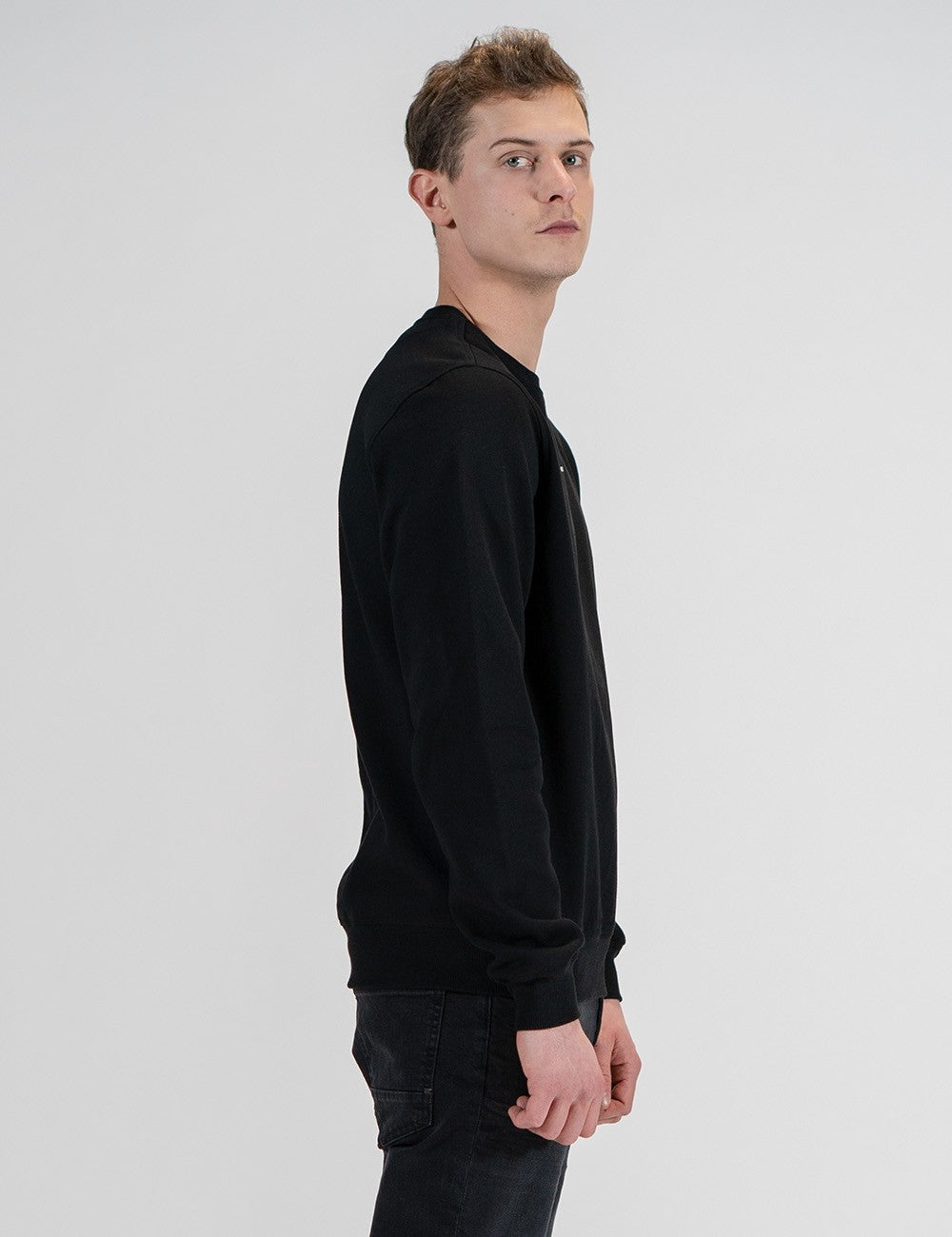 Side profile of a man with short brown hair modeling a long-sleeved, crewneck black sweatshirt and dark jeans against a plain light gray background.