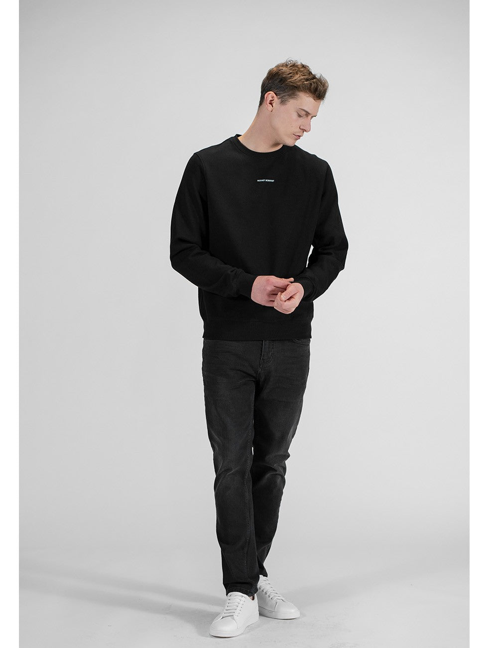 A man stands looking down, wearing a black long-sleeve crew-neck sweatshirt featuring a small white logo on the center chest. He is dressed in dark grey slim-fit jeans paired with clean white low-top sneakers against a plain light grey background.