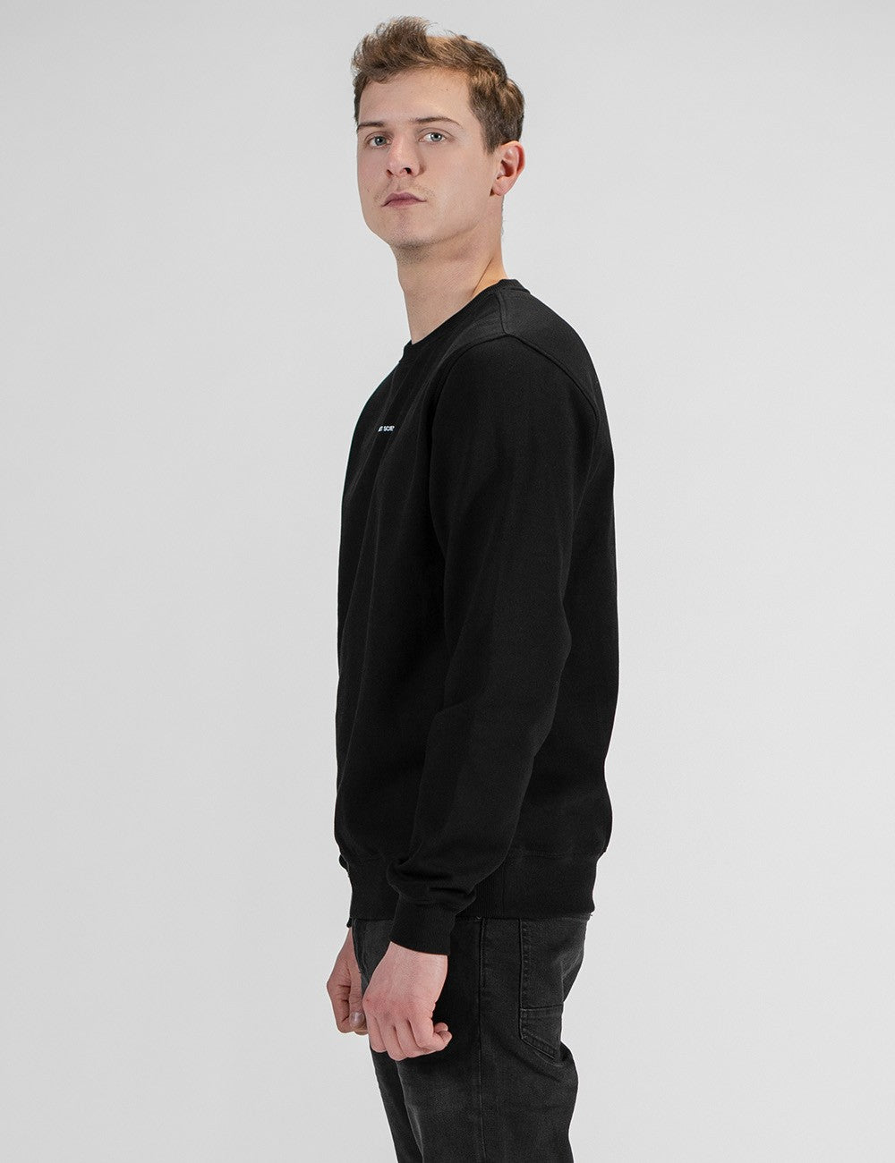 Side profile of a man with short brown hair modeling a long-sleeved, crewneck black sweatshirt and dark jeans against a plain light gray background.