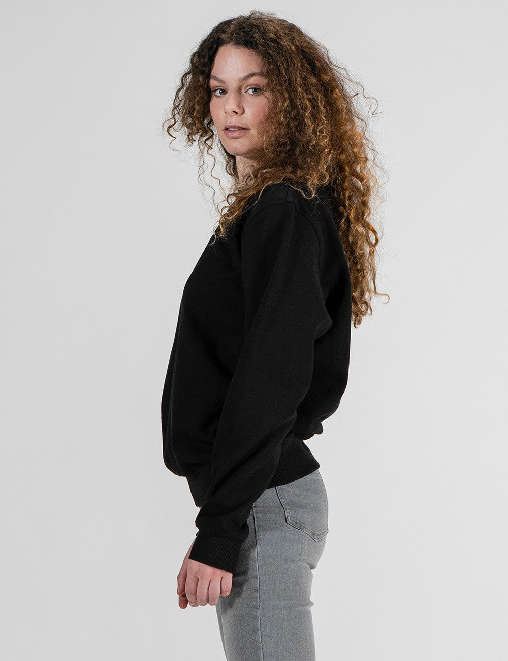 A side-angle view of a woman with long, voluminous, curly brown hair wearing a solid black crewneck sweatshirt and grey pants against a plain white background.