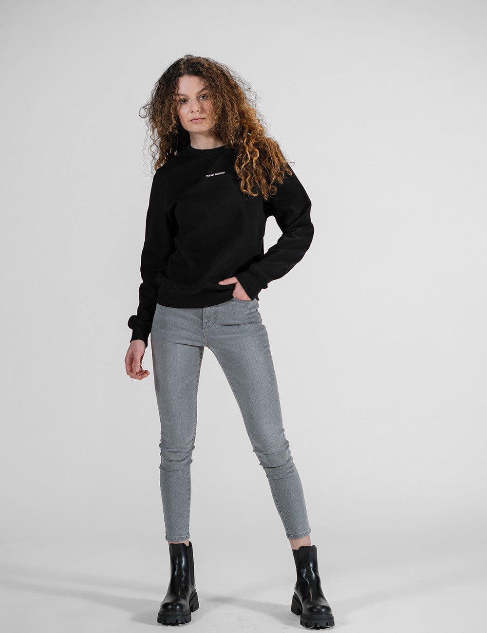 A young woman with long, voluminous, curly brown hair stands facing forward against a plain light gray background. She is wearing a black long-sleeved crew-neck sweatshirt featuring a small white graphic text across the center chest. Her outfit includes gray skinny jeans and a pair of black chunky ankle boots with thick, lugged soles. She has her left hand resting casually in her pants pocket while her right arm hangs loose at her side.