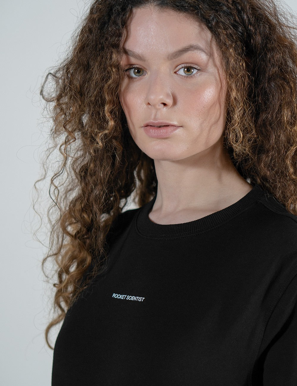 A close-up portrait of a woman with long, voluminous, curly brown hair looking directly into the camera. She has a fair complexion, subtle makeup, and a small nose piercing. She is wearing a plain black t-shirt featuring the words "ROCKET SCIENTIST" printed in a small, white, minimalist sans-serif font across the upper chest.