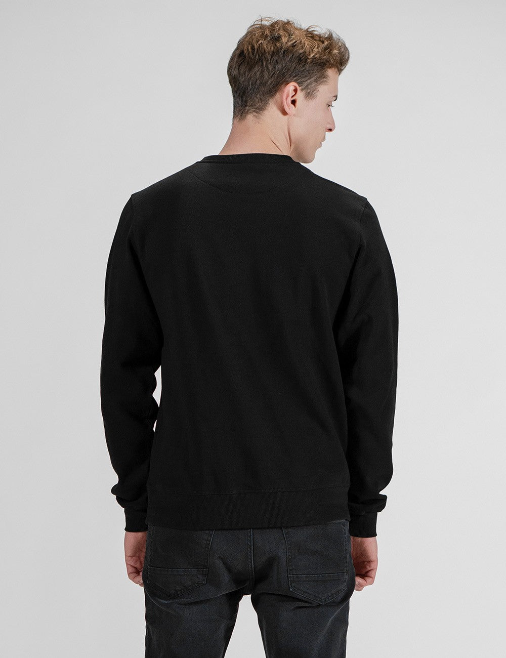 A man with short, wavy blonde hair is shown from behind, wearing a plain black long-sleeve sweatshirt and dark grey or black jeans against a light grey background.