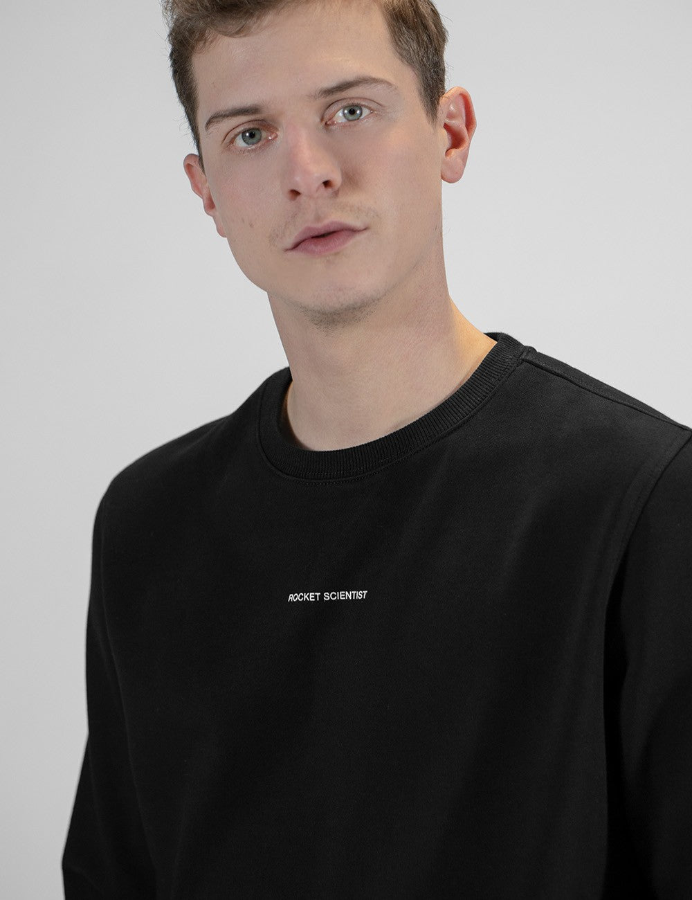 A young man with light brown hair, fair skin, and blue eyes looking directly forward. He is wearing a black crewneck sweatshirt or long-sleeve shirt. On the center of the chest, there is a small, minimalist text in white all-caps that reads "ROCKET SCIENTIST".