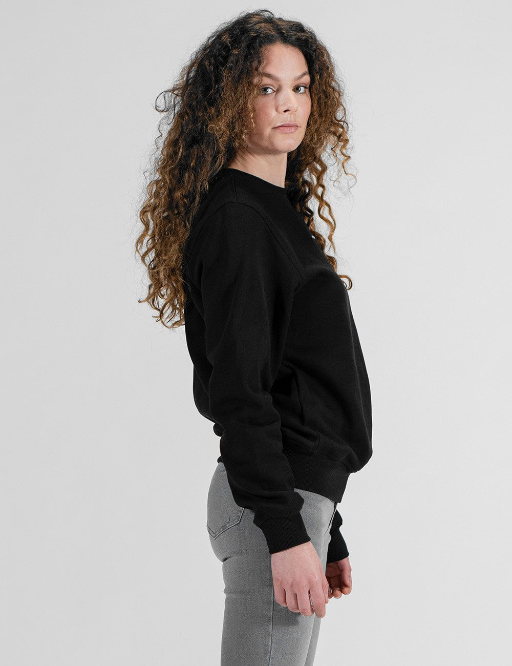 A side-angle view of a woman with long, voluminous, curly brown hair wearing a solid black crewneck sweatshirt and grey pants against a plain white background.